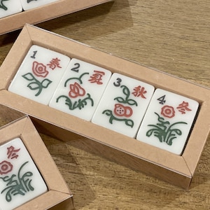 May include: Four white tiles with red and green floral designs and Chinese characters. The tiles are arranged in a row in a clear plastic case with a brown cardboard backing. The tiles are numbered 1 through 4.