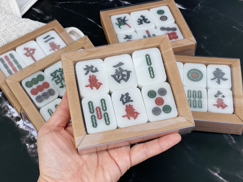 May include: A set of white mahjong tiles with black and red Chinese characters and green and red symbols. The tiles are arranged in a rectangular box with a clear plastic lid and a brown cardboard base.