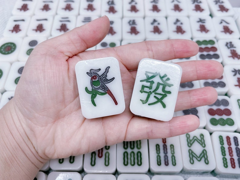 May include: Two white mahjong tiles with green and black designs. One tile features a bird with red accents and the other tile has the Chinese character for "East" in green.