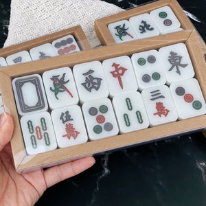 May include: A set of white mahjong tiles with black and green Chinese characters and red and green dots. The tiles are arranged in a rectangular box with a brown cardboard exterior.