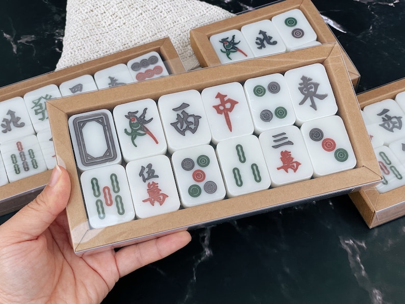 May include: A set of white mahjong tiles with black and green Chinese characters and red and green dots. The tiles are arranged in a rectangular box with a brown cardboard exterior.