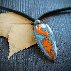 Könnte beinhalten: Ein blauer und orangefarbener Emaille-Anhänger mit einem Fuchs-Design hängt an einem schwarzen Lederband. Der Anhänger liegt auf einer dunklen Holzoberfläche mit einem getrockneten Blatt.