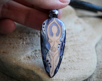 May include: A ceramic pendant necklace with a blue and white design featuring a stylised female figure. The pendant is shaped like a teardrop and has a black leather cord.