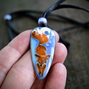 Könnte beinhalten: Eine Halskette mit einem blauen und orangefarbenen Emaille-Anhänger, der einen Fuchs mit eingerolltem Schwanz zeigt. Der Anhänger ist tropfenförmig und hat eine schwarze Lederschnur.
