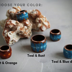 May include: Four ceramic rings in different color combinations: teal and rust, rust and orange, teal and blue dots, and teal and rust. The rings are displayed on a white surface with a piece of coral.