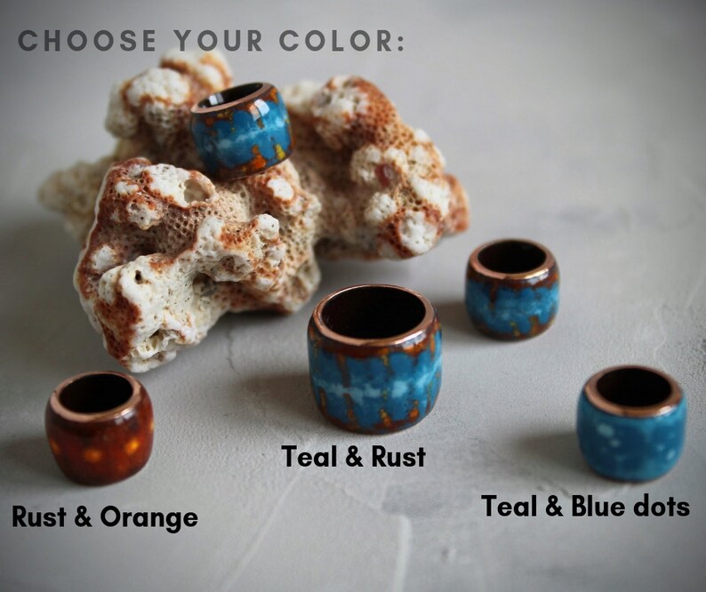 May include: Four ceramic rings in different color combinations: teal and rust, rust and orange, teal and blue dots, and teal and rust. The rings are displayed on a white surface with a piece of coral.