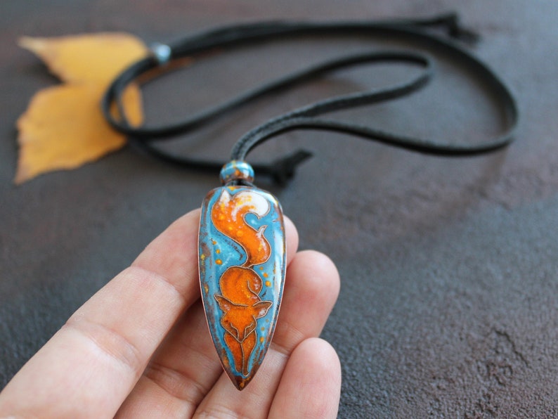 May include: A pendant necklace with a blue and orange fox design. The pendant is shaped like a teardrop and has a black cord.