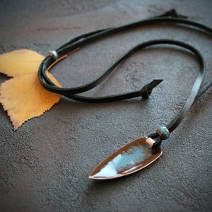 May include: A black leather cord necklace with a copper pendant featuring a blue and brown glaze. The pendant is shaped like a teardrop and has a pointed tip.