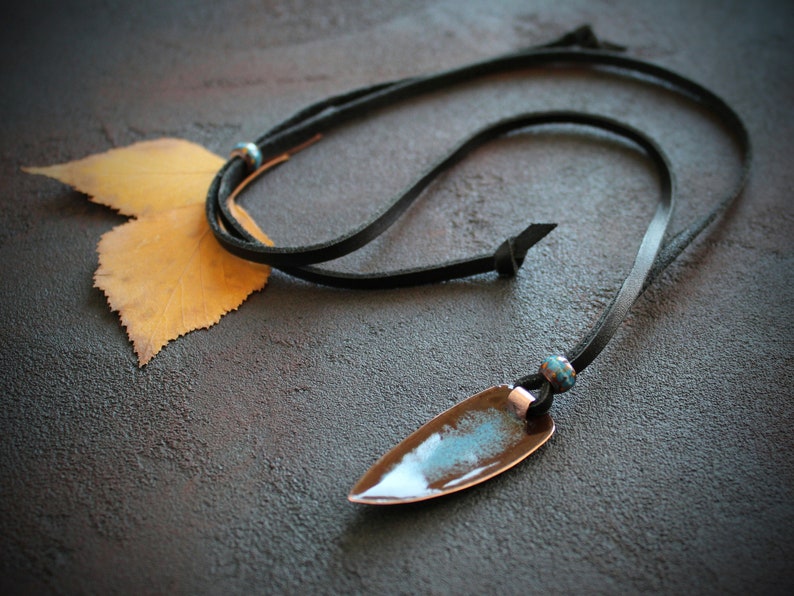 May include: A black leather cord necklace with a copper pendant featuring a blue and brown glaze. The pendant is shaped like a teardrop and has a pointed tip.
