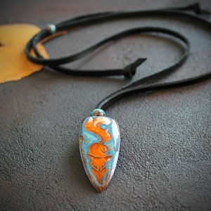 May include: A blue and orange fox pendant necklace on a black leather cord. The pendant is shaped like a teardrop and has a detailed fox design.
