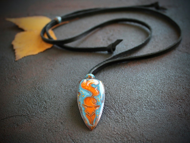 May include: A blue and orange fox pendant necklace on a black leather cord. The pendant is shaped like a teardrop and has a detailed fox design.