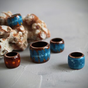 May include: Five ceramic rings with a copper base and blue and orange glaze. The rings are arranged on a white surface with a piece of coral behind them.