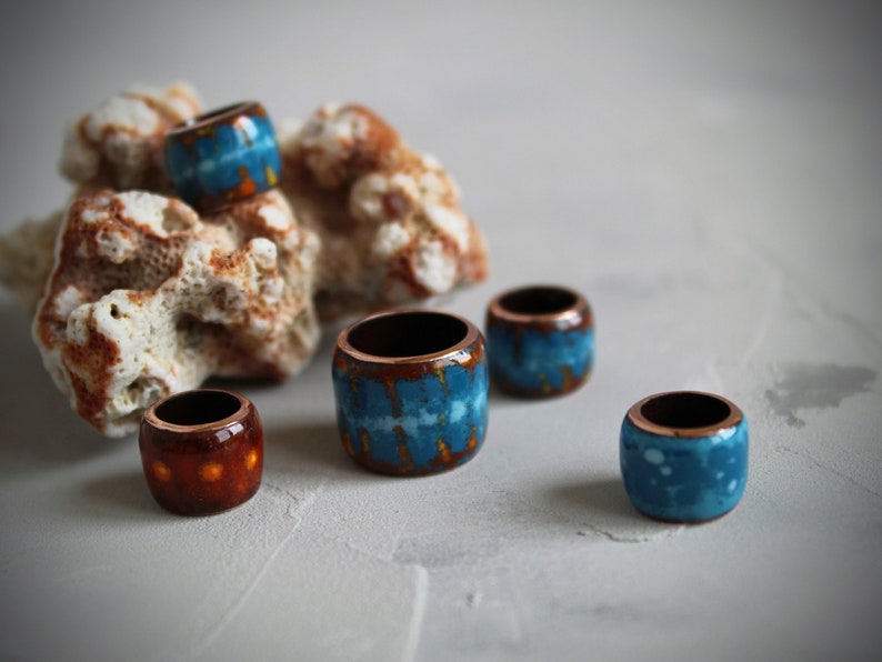 May include: Five ceramic rings with a copper base and blue and orange glaze. The rings are arranged on a white surface with a piece of coral behind them.