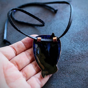 K&ouml;nnte beinhalten: Eine Halskette aus schwarzem Lederband mit einem blauen und schwarzen Emaille-Anh&auml;nger. Der Anh&auml;nger ist fischf&ouml;rmig und hat kupferfarbene Akzente.