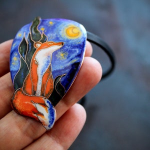 Puede incluir: Un colgante de zorro con un diseño de esmalte azul, naranja y verde. El colgante tiene forma de media luna y presenta un cielo nocturno estrellado con una luna creciente.