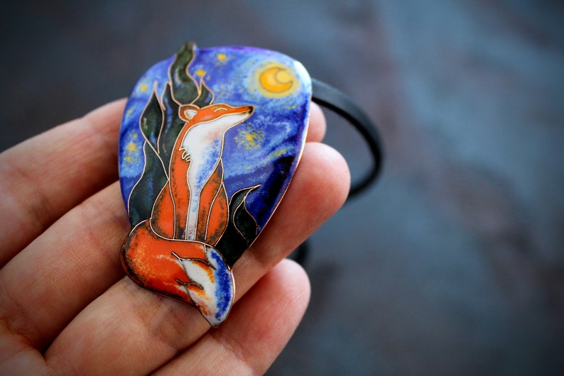 K&ouml;nnte beinhalten: Ein Fuchs-Anh&auml;nger mit einem blauen, orangefarbenen und gr&uuml;nen Emaille-Design. Der Anh&auml;nger ist in Form eines Halbmondes geformt und zeigt einen sternenklaren Nachthimmel mit einem Halbmond.