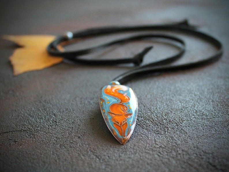 May include: A blue and orange enamel pendant with a fox design, hanging from a black leather cord.