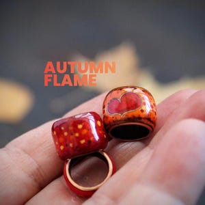 Puede incluir: Dos anillos cilíndricos pequeños con un acabado esmaltado brillante en tonos rojo, naranja y amarillo. Un anillo presenta un diseño de corazón rojo prominente, mientras que el otro tiene un patrón moteado. El texto "AUTUMN FLAME" es visible.