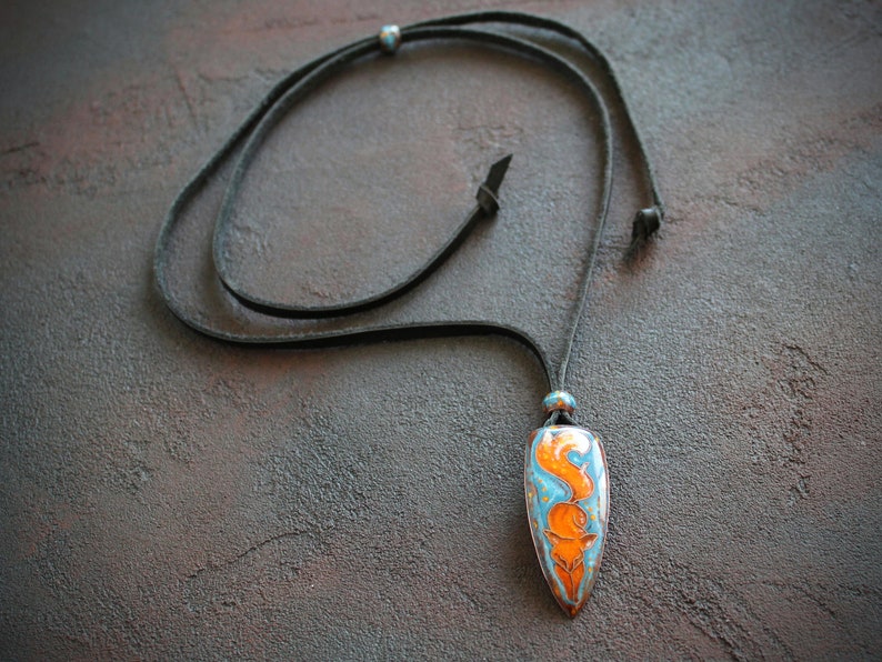 May include: A necklace with a leather cord and a pendant shaped like a teardrop. The pendant is made of ceramic and has a blue and orange design.