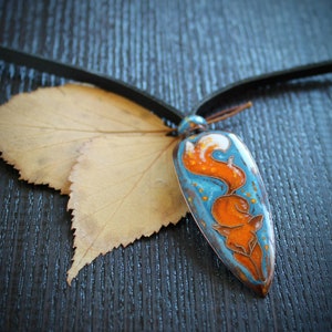May include: A blue and orange enamel pendant with a fox design hangs from a brown leather cord. The pendant is shaped like a teardrop and has a white background with a blue and orange fox design. The fox is curled up in a ball with its tail wrapped around its body. The pendant is resting on a brown wooden surface.