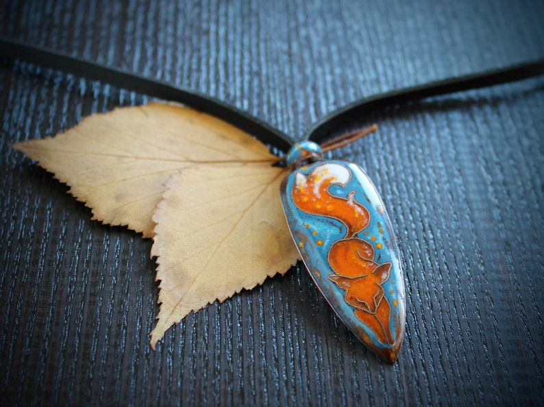 May include: A blue and orange enamel pendant with a fox design hangs from a brown leather cord. The pendant is shaped like a teardrop and has a white background with a blue and orange fox design. The fox is curled up in a ball with its tail wrapped around its body. The pendant is resting on a brown wooden surface.