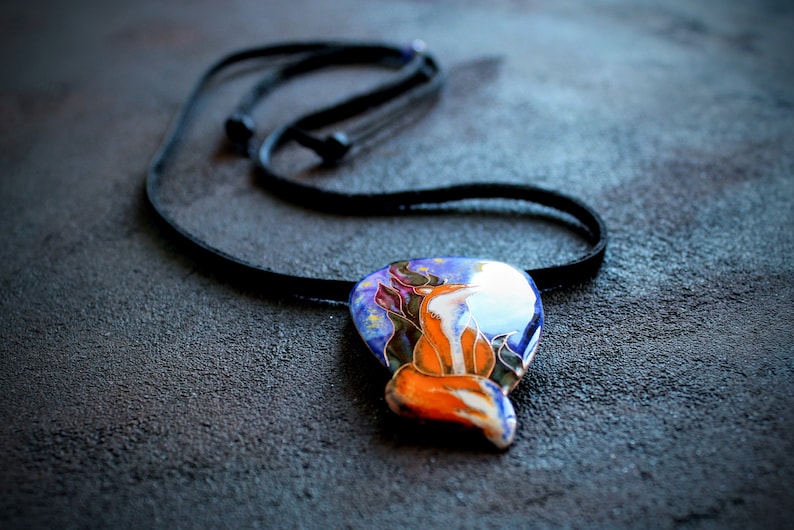 K&ouml;nnte beinhalten: Eine Halskette mit einem blauen, orangefarbenen und wei&szlig;en Anh&auml;nger, der ein stilisiertes Fuchsdesign zeigt. Der Anh&auml;nger ist vor einem Hintergrund aus Sternen und einem dunkelblauen Himmel platziert.