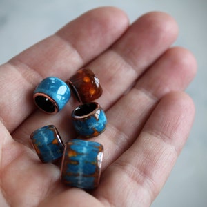 May include: A collection of five colorful ceramic beads with copper accents. The beads are in shades of blue and orange with white marbling.