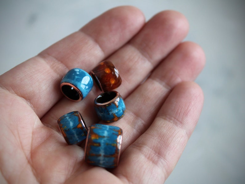 May include: A collection of five colorful ceramic beads with copper accents. The beads are in shades of blue and orange with white marbling.