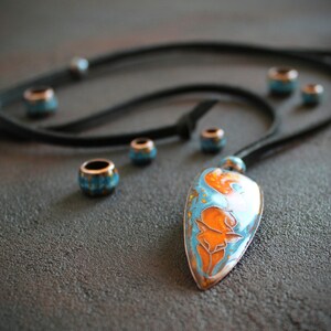 May include: A turquoise and orange enamel pendant with a floral design hangs from a black leather cord with turquoise and copper beads.
