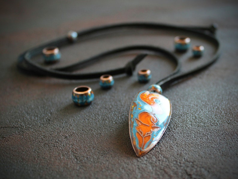 May include: A turquoise and orange enamel pendant with a floral design hangs from a black leather cord with turquoise and copper beads.