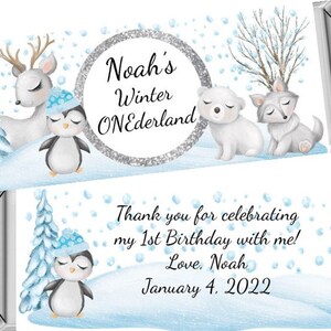 May include: A blue and white winter wonderland themed chocolate bar wrapper with a penguin, deer, polar bear, and wolf. The wrapper says "Noah's Winter ONEderland" and "Thank you for celebrating my 1st Birthday with me! Love, Noah January 4, 2022".