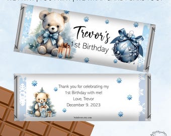 May include: Two candy bar wrappers featuring a winter wonderland theme with light blue snowflakes, a teddy bear with a blue bow, and a blue ornament. The text reads: BOY'S BIRTHDAY WINTER WONDERLAND, WE PRINT | YOU PRINT | WE WRAP CANDY BARS TOO!, Trevor's 1st Birthday, and Thank you for celebrating my 1st Birthday with me! Love, Trevor December 9. 2023.