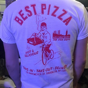 May include: White t-shirt with red text that says "BEST PIZZA" in a curved arc. The shirt features a cartoon illustration of a person riding a bicycle with a pizza box on the back. The text also says "HOT & DELICIOUS", "WILLIAMSBURG 718 599 2210", "DINE-IN TAKE-OUT DELIVERY", and "33 HAVEMEYER ST. BROOKLYN, NY 11211".