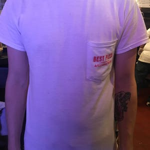 May include: White t-shirt with a red pocket that says "Best Pizza Williamsburg" in red text.