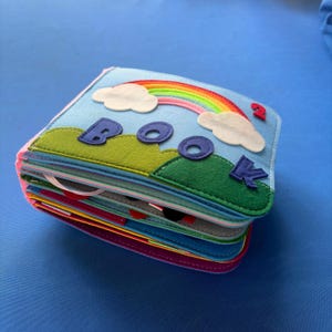 May include: A multi-layered felt quiet book with a light blue cover featuring a rainbow, white clouds, green hills, and the purple felt letters spelling BOOK. The number 2 is stitched in red felt on the top right corner of the cover.