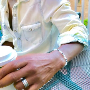 May include: A white beaded bracelet with black accents on a person's wrist. The bracelet is made of small, round beads and has a simple design.