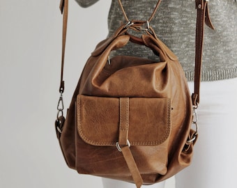 Può includere: Uno zaino in pelle marrone con una tasca frontale e spallacci regolabili. La borsa ha una forma arrotondata e una maniglia superiore. Gli spallacci sono attaccati con hardware color argento. Lo zaino è mostrato su uno sfondo neutro.