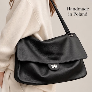Puede incluir: Un bolso de hombro negro de cuero con superficie texturizada, solapa plegable y cierre giratorio de metal plateado. El bolso cuenta con una correa larga para el hombro e incluye el texto Handmade in Poland impreso en la parte superior derecha.