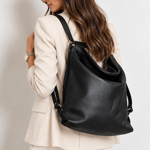Puede incluir: Una mochila de cuero negro con textura granulada, que presenta una silueta holgada y desestructurada, correas finas para los hombros y herrajes de metal en tono dorado. El bolso tiene un compartimento principal grande con diseño plegable.