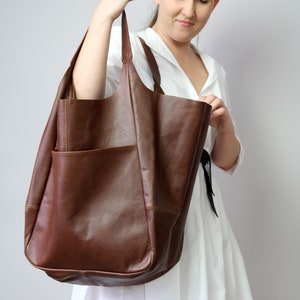 May include: A brown leather tote bag with a large pocket on the front. The bag is being held by a person wearing a white dress.