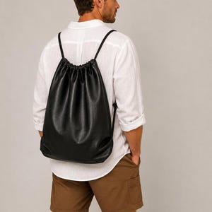 May include: A black drawstring backpack made of a smooth, matte material. The bag features a gathered top closure with thin black cord straps that extend from the top to the bottom corners, creating a simple and functional design for carrying items.