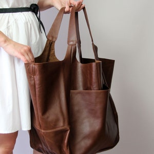 May include: A large brown leather tote bag with a spacious interior and two large pockets on the front. The bag has a wide opening and two sturdy handles.