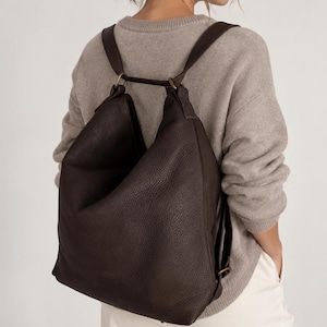 Könnte beinhalten: Ein dunkelbrauner Lederrucksack mit genarbter Textur, der eine lockere, unstrukturierte Silhouette aufweist, mit zwei Schultergurten und Metallringen in antikem Messington, die die Gurte mit der Oberseite der Tasche verbinden.