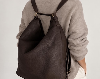 May include: A dark brown leather backpack with a pebbled texture, featuring a slouchy, unstructured silhouette, two shoulder straps, and antique brass-toned metal hardware rings connecting the straps to the top of the bag.