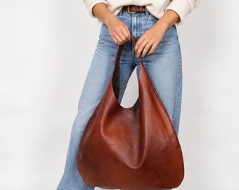 May include: A large, slouchy hobo-style shoulder bag crafted from smooth, deep brown leather. The bag features a wide, integrated shoulder strap and a spacious, unstructured main compartment with a dark interior lining, showcasing a minimalist design.