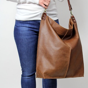 Puede incluir: Un bolso tote de cuero marr&oacute;n con una correa larga. El bolso se lleva sobre el hombro de una persona que lleva una camisa blanca de manga larga y pantalones vaqueros azules.