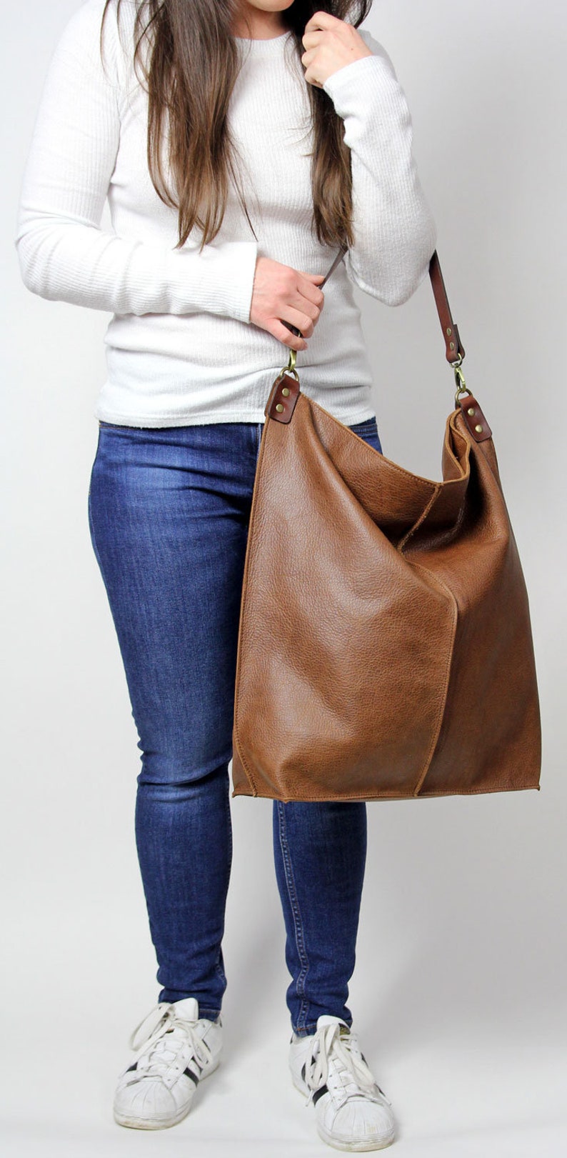 Puede incluir: Un bolso tote de cuero marr&oacute;n con una correa larga. El bolso se lleva sobre el hombro de una persona que lleva una camisa blanca de manga larga y pantalones vaqueros azules.