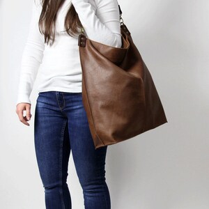 Puede incluir: Una mujer con una camisa blanca de manga larga, jeans azules y zapatillas blancas lleva un bolso tote de cuero marr&oacute;n grande.