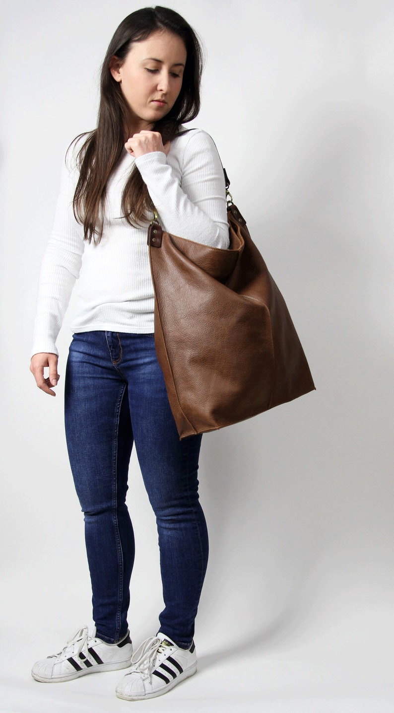 Puede incluir: Una mujer con una camisa blanca de manga larga, jeans azules y zapatillas blancas lleva un bolso tote de cuero marr&oacute;n grande.