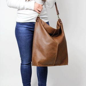Puede incluir: Un bolso tote de cuero marr&oacute;n con una correa larga. El bolso es grande y espacioso, y lo lleva una persona que lleva jeans y una camisa blanca.